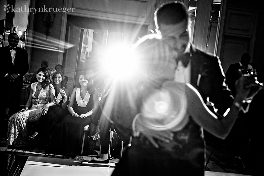 Black and white of groom and mother dancing with bride looking on.