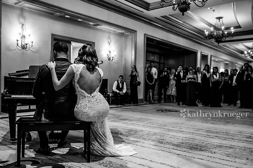 Black and white of bride and groom sitting at piano bench with guests looking on.