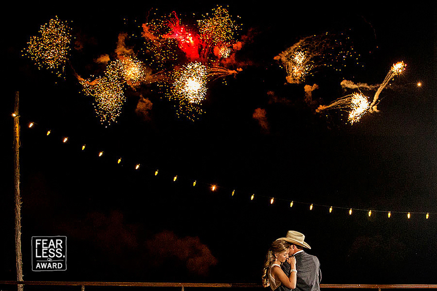 Bride and groom embrace with fireworks overhead.